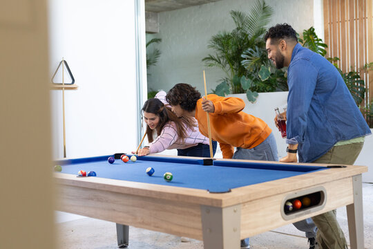 Diverse friends leaning and lining up shots at game room with blue-felt pool table and cues