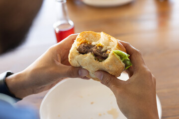 Mid-adult man holding half-eaten burger over plate on wooden table, red bottle, wristwatch showing