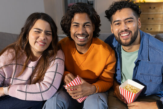 Friends sharing red-and-white striped popcorn boxes and laughing on gray sofa in cozy living room