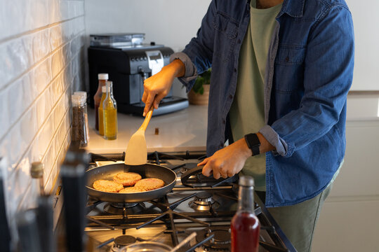 Cook is flipping four burger patties on gas stovetop using spatula beside oil bottles, coffee maker