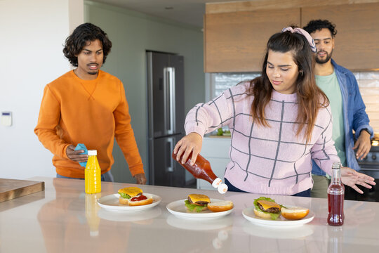 Diverse friends pouring ketchup on burgers at island countertop in kitchen with blue cleaning cloth