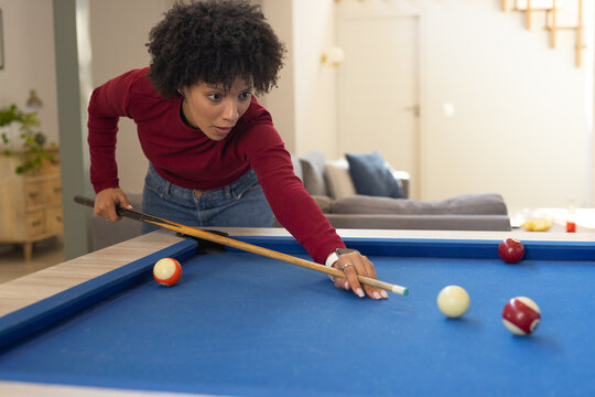 African American woman leaning over blue pool table in living room aiming wooden cue