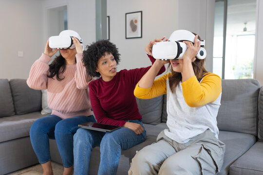 Diverse female friends sitting on gray sofa in living room using VR headsets holding tablet - Powered by Adobe