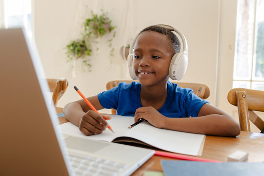 African American boy sitting at table wearing headphones writing with orange pencil checking laptop
