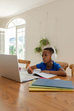 African American child sitting at wooden dining table at home, working on laptop with orange pencil