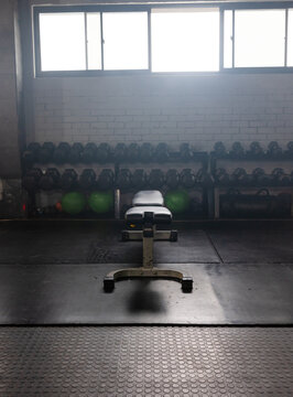 Adjustable workout bench is sitting in dim gym weight area, with dumbbell rack and kettlebells