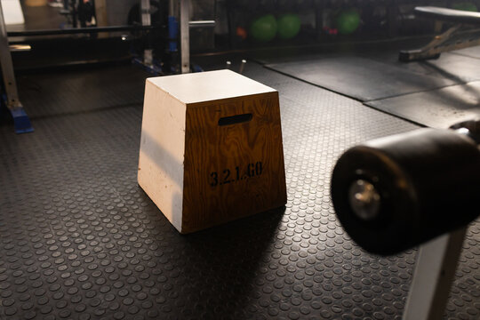 Wooden plyo box is sitting on coin-pattern rubber floor in gym, casting long warm shadow