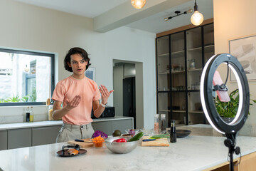 Asian man filming cooking, mixing vegetables in metal bowl with smartphone ring light in kitchen