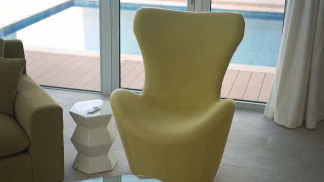 sunlit yellow chair by pool window modern interior lounge with textured upholstery, white geometric side table, beige sofa arm, flowing curtains, glass doors