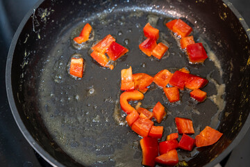 Chopped red bell peppers cooking in a sizzling frying pan with oil. The vibrant colors of the peppers contrast against the dark surface, indicating a healthy meal preparation.