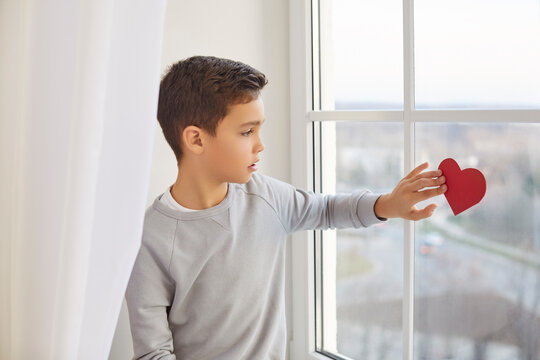 Lonely little boy touching red paper heart on glass looking out window, staying at home alone, child waiting for parents thinking about school problems, bad relationship in family, psychological worry