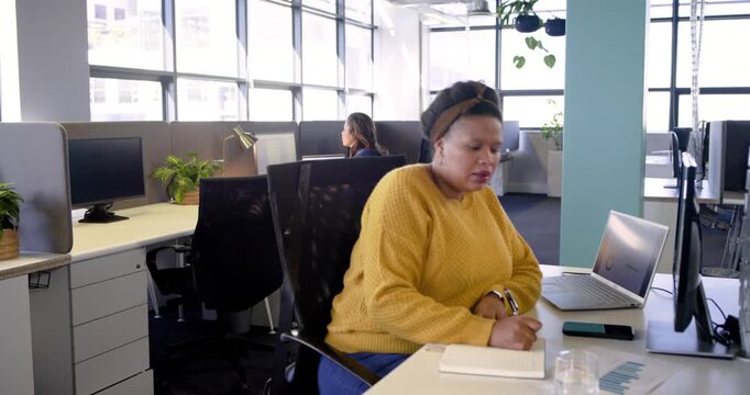 African American woman facing laptop bug, syncing smartphone, noting and reviewing report at office