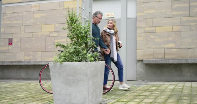 Senior couple pushing bicycle sipping coffee in plaza hooking helmet admiring facade while laughing