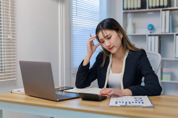 Stressed businesswoman feeling tired and having a headache while using a calculator and working on a laptop in an office