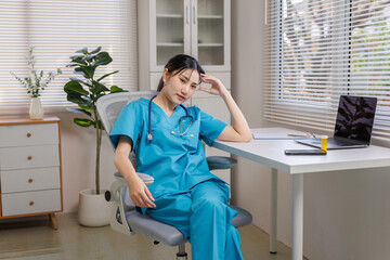 Female healthcare worker in scrubs looking exhausted and stressed, sitting at a desk in an office,...