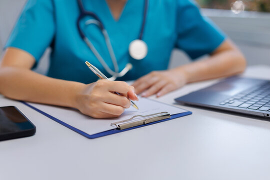 Healthcare worker wearing scrubs and stethoscope, writing important medical information on a clipboard at a desk in a hospital or clinic