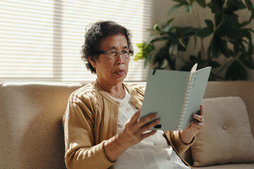 Senior Asian woman relaxing on a sofa, wearing glasses and reading a notebook by sunlight. Enjoying leisure time