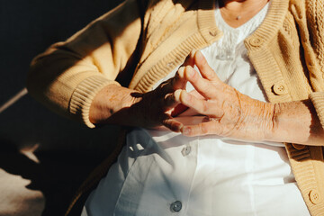 Naklejka premium Elderly woman with clasped, wrinkled hands resting. Sunlight illuminating her creating strong shadows. Showing aging, serenity, and contemplation