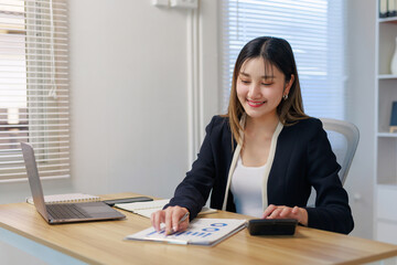 Young Asian businesswoman using calculator and report, analyzing data for financial planning and smiling in a modern office