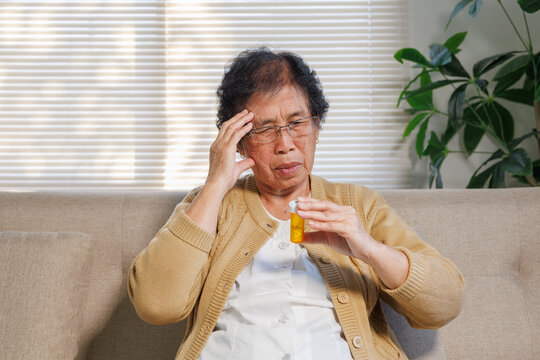 Senior woman suffering from head pain and confusion while holding a pill bottle, experiencing health issues at home