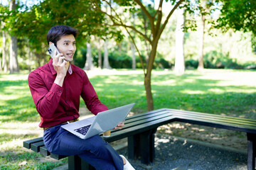 Professional man talks on phone at park while working on laptop during sunny day
