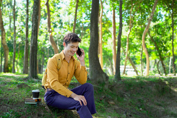 Fototapeta premium Happy asian man using a smartphone in a park, enjoying a pleasant conversation on a sunny day, surrounded by lush greenery and modern architecture.