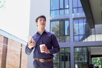 Smiling businessman using smartphone and holding coffee outside office building