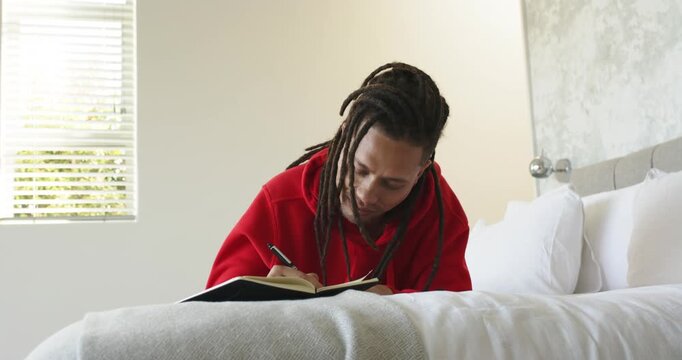 Glancing toward window and gathering thoughts, man writing with pen in notebook on bed in bedroom