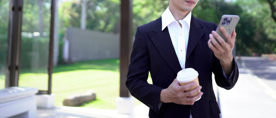 Young asian businessman in black suit with takeout coffee using smartphone at urban park, blank space