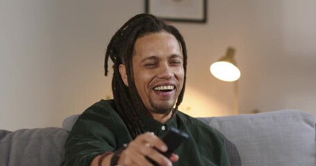 Mid adult African American man clicking remote for fun on gray sofa under brass lamp, smiling
