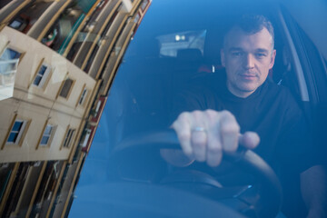 Male driver looking ahead while sitting inside a car, visible through front windshield with building reflection