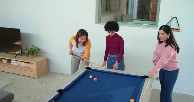 Diverse female friends lining up shot at rec room pool table, striking cue ball and celebrating