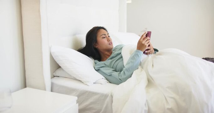 Feeling tired, mid adult Asian woman in bed scrolling purple phone, pulling duvet and closing eyes
