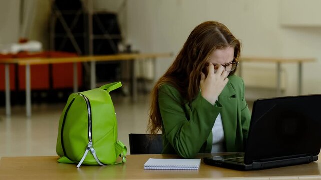 Exhausted college girl in bright green jacket covers her face with hands in front of laptop during study session, concept deadline pressure and academic anxiety.