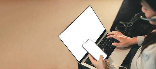 Asian woman wearing headset working from home with laptop and smartphone. Remote 24/7 customer service support concept with blank screens for UI mockup, communication, and digital workspace.