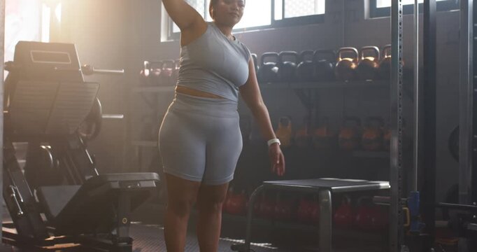 After brief pause mid adult woman shifting weight onto left leg leaning right amid gym equipment