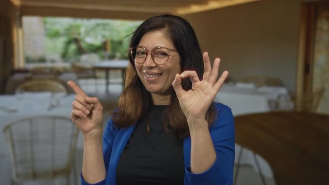 Woman in blue jacket making ok sign with her right hand and pointing left index finger in restaurant dining area; approval.