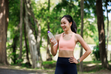Fototapeta premium Young Asian woman smiling, holding a water bottle, and staying hydrated during an outdoor exercise session in a park