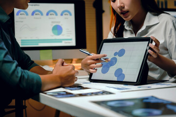 Business colleagues discussing corporate financial report data using a digital tablet and computer monitor during a late night meeting