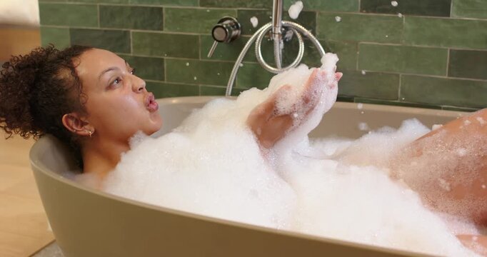 Foam on chest, African American woman reclining in tub, scooping and blowing bubbles, relaxing