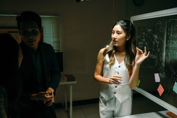 Young Asian business woman presenting ideas on a whiteboard during a team meeting in a dark modern office environment