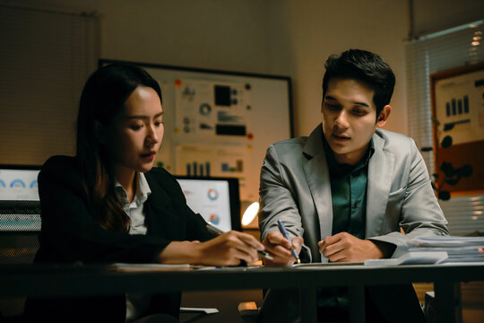 Two diligent business colleagues collaborating on a project, analyzing data and discussing strategy while working late at night in an office - Powered by Adobe