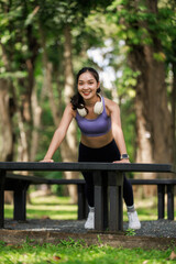 Young Asian woman performing push ups using a park bench, staying active and focused on her workout with a smile