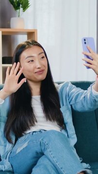 Asian woman in casual clothes using smartphone sitting on the couch at home and enjoying her video call with friend. Leisure concept.