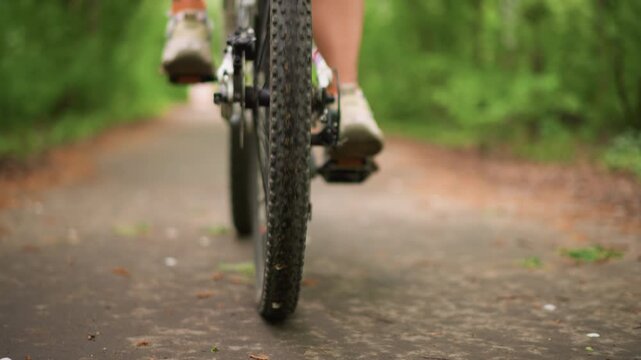 CloseUp Front Wheel On Forest Path, White Woman Pedals Along Leafy Asphalt Lane, Textured Tire Treads And Spinning Spokes, Soft Bokeh Green Canopy, Steady LowAngle Motion For Sports Wellness Footage