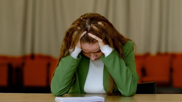 Young student with long hair in green jacket leans forward and grabs her head in frustration, cannot learn material before difficult test, concept cramming overload and academic anxiety.