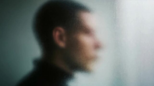 Abstract view of man in profile behind fogged glass gives moody cinematic feeling of introspection mystery and emotional distance suitable for mental health or storytelling concepts