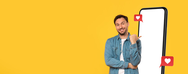 A man stands in front of a bright yellow background, smiling and pointing to a large, blank smartphone. The scene creates an energetic vibe, perfect for promoting digital engagement and connection.