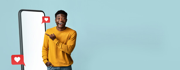 A smiling man points at a large phone in a light blue background. The display features heart icons, representing positive social media engagement, creating an energetic atmosphere.