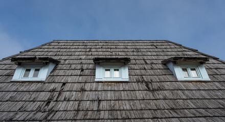 A dramatic low-angle view of a rustic wooden roof, showing faded wooden planks arranged in strong geometric lines. Three blue windows are visible on the roof.